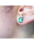 Oval natural emerald and diamond earrings in 14K white gold on an ear.
