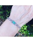 A paperclip gold bracelet with a real emerald on a wrist against a natural background.