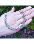 Hand displaying a white gold and emerald molecule-shaped bracelet with a blurred green outdoor background.