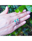 Hand holding a pair of natural teardrop emerald earrings with a leafy background