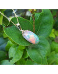 Ethiopian Welo opal necklace pendant with red and rainbow fire in 14k yellow gold, resting on green leaves.