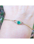 Gold bracelet with a square emerald on a wrist against a blurred natural background.