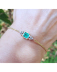 Gold bracelet with a emerald-cut emerald on a wrist against a blurred natural background.