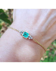 Gold bracelet with a emerald-cut emerald on a wrist against a blurred natural background.