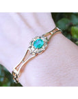 Gold bracelet with an oval cut natural emerald on a wrist against a blurred green background.