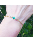 Gold bracelet with a rectangular emerald on a wrist against a blurred green background.
