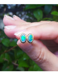 Opal stud earrings held between fingers with a blurred green leafy background