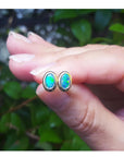 Opal stud earrings held between fingers with a blurred green leafy background