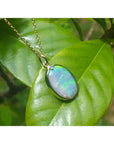Oval Australian opal pendant with green-bluish rainbow play, set in 14K yellow gold, resting on a green leaf.