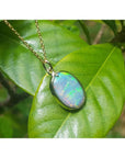 Oval Australian opal pendant with green-bluish rainbow play, set in 14K yellow gold, resting on a green leaf.