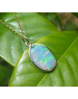 Oval Australian opal pendant with green-bluish rainbow play, set in 14K yellow gold on a green leaf.