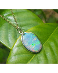 Oval Australian opal pendant with green-bluish rainbow play, set in 14K yellow gold on a green leaf.