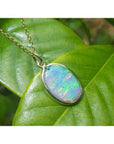 Oval Australian opal pendant with green-bluish rainbow play, set in 14K yellow gold on a green leaf.
