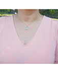 A woman wearing a pink top with a necklace featuring a blue opal pendant outdoors.