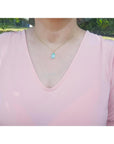 A woman wearing a pink top with a necklace featuring a blue opal pendant outdoors.