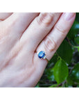 Hand wearing a ring with a blue sapphire against a green leafy background