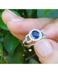 14k white gold ring with a blue oval cut natural sapphire held between fingers against a green leafy background