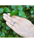 Hand wearing a 14k white gold ring with a blue natural sapphire oval cut against a green leafy background