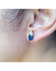 Oval blue sapphire stud earring with three small diamonds, set in 14K white gold on an ear.