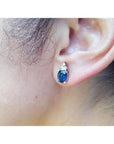 Oval blue sapphire stud earring with three small diamonds, set in 14K white gold on an ear.