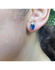 Oval blue sapphire stud earrings with three prong-set diamonds in 14K white gold on an ear.