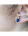 An ear wearing a blue oval cut sapphire gemstone earring topped with three prong-set natural diamonds.