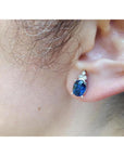An ear wearing a blue oval cut sapphire gemstone earring topped with three prong-set natural diamonds.