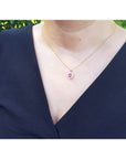 Necklace with a pink oval cut sapphire worn by a woman in a black top, with a blurred green background.