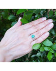 Hand wearing an oval cut emerald ring with accent diamonds against a green leafy background