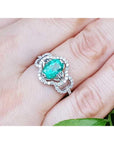 A 14k white gold clover ring with an oval cut emerald surrounded with natural diamonds on a person's finger.