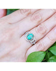 A clover ring with an oval cut emerald on a hand against a green leafy background.