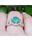 A white gold emerald ring on a finger against a blurred green leafy background.