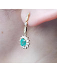 Oval emerald and halo diamond dangle earrings in 14K yellow gold, worn on an ear.