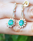 Gold earrings with emerald stones held in a hand against a blurred green background