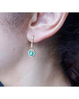 Oval emerald and halo diamond dangle earrings in 14K yellow gold on an ear.
