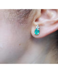 Oval cut natural emerald earrings with three diamonds in 14K white gold on an ear.