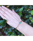 Hand wearing a gold bracelet with a natural emerald against a green leafy background.