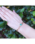 Hand wearing a gold bracelet with a natural emerald against a green leafy background.