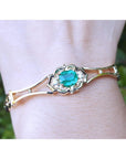 Gold bracelet with a natural colombian emerald oval cut on a wrist against a blurred green background.
