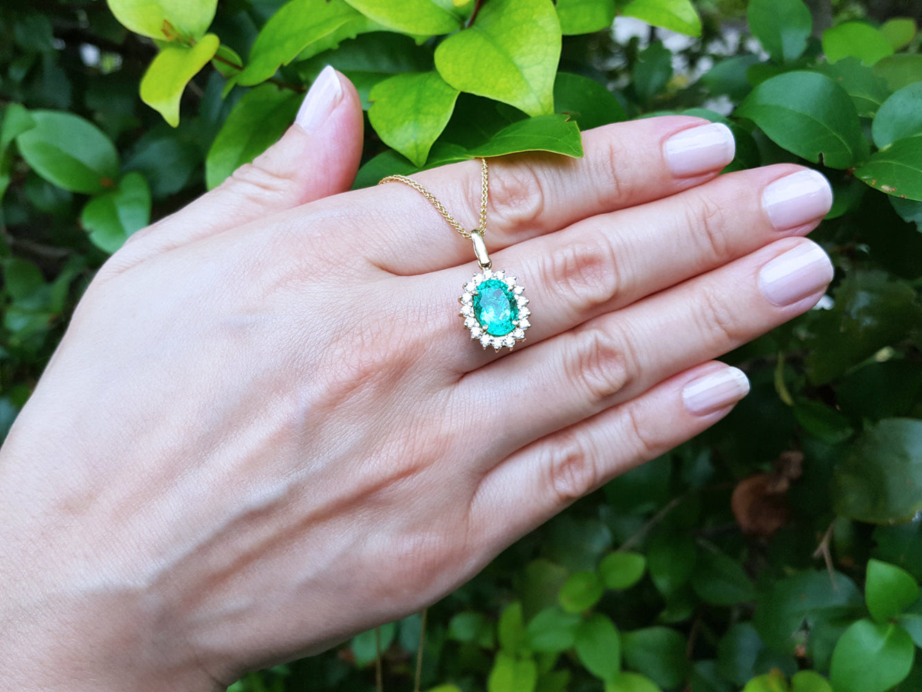Hand displaying a gold necklace with an emerald pendant against a green leafy background.