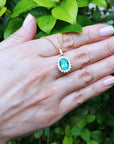 Hand displaying a gold necklace with an emerald pendant against a green leafy background.