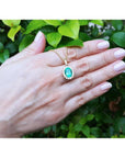 Oval natural emerald pendant with halo diamonds in 18K yellow gold, worn on a hand.