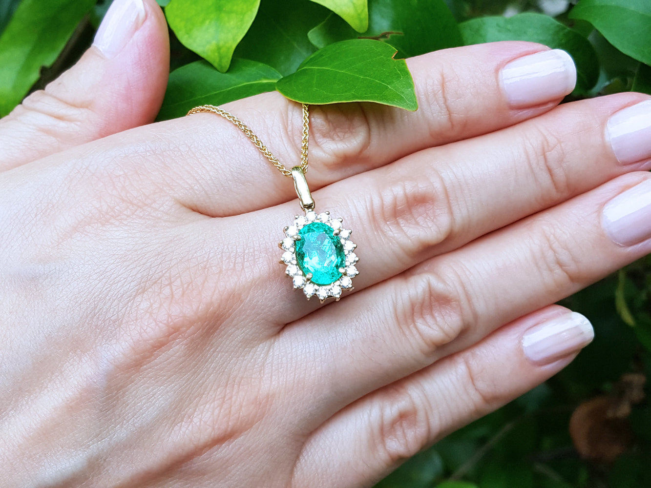 Gold necklace with a natural emerald pendant on a hand against a green leafy background.