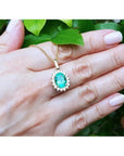 Oval natural emerald pendant with halo diamonds in 18K yellow gold, held in a hand.