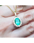 Oval natural emerald pendant with halo diamonds in 18K yellow gold, worn on a hand.