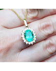 Oval natural emerald pendant with halo diamonds in 18K yellow gold, worn on a hand.