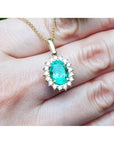 Oval natural emerald pendant with halo diamonds in 18K yellow gold, worn on a hand.