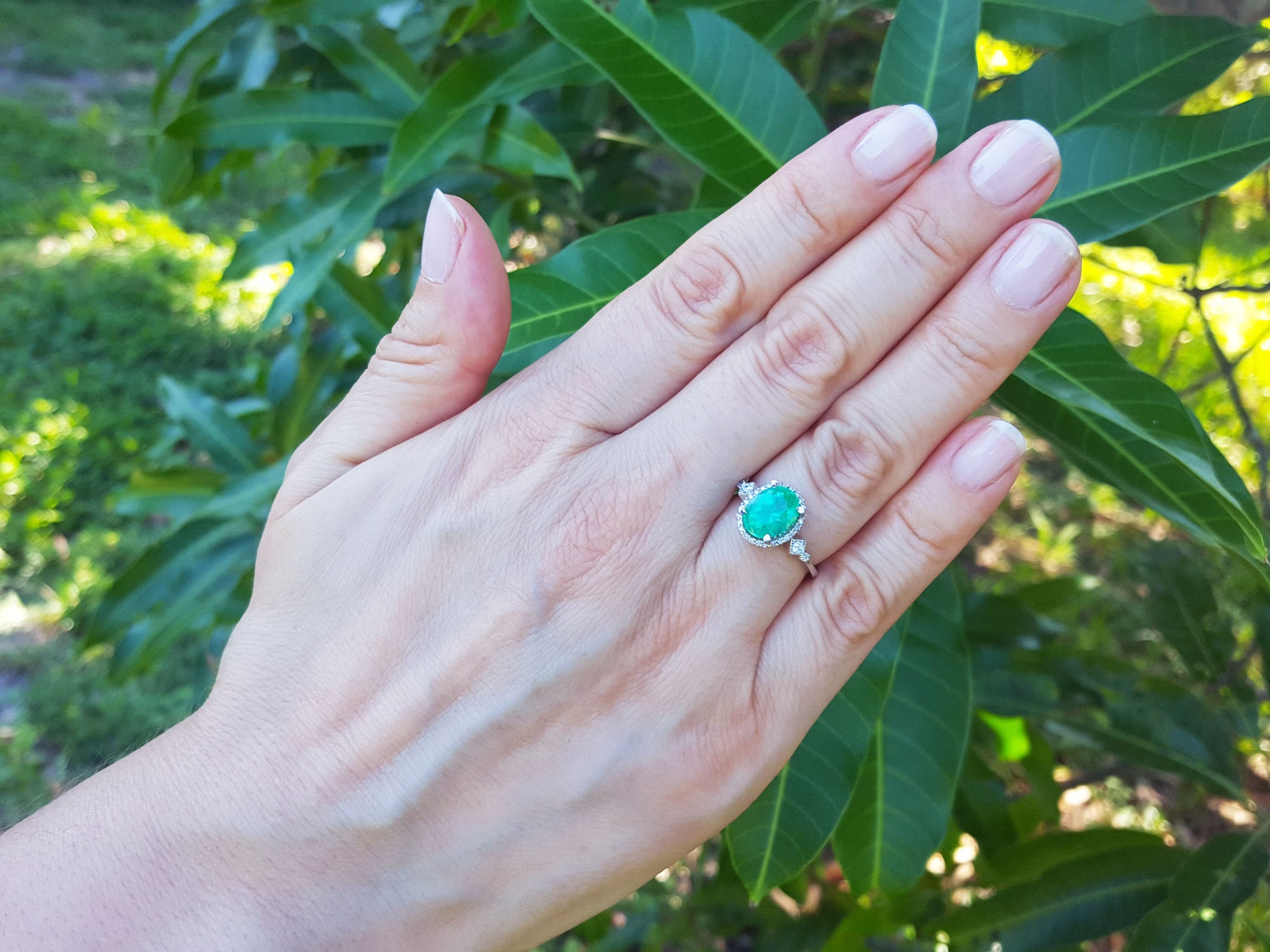 Hand wearing an oval emerald ring with leaves in the background,