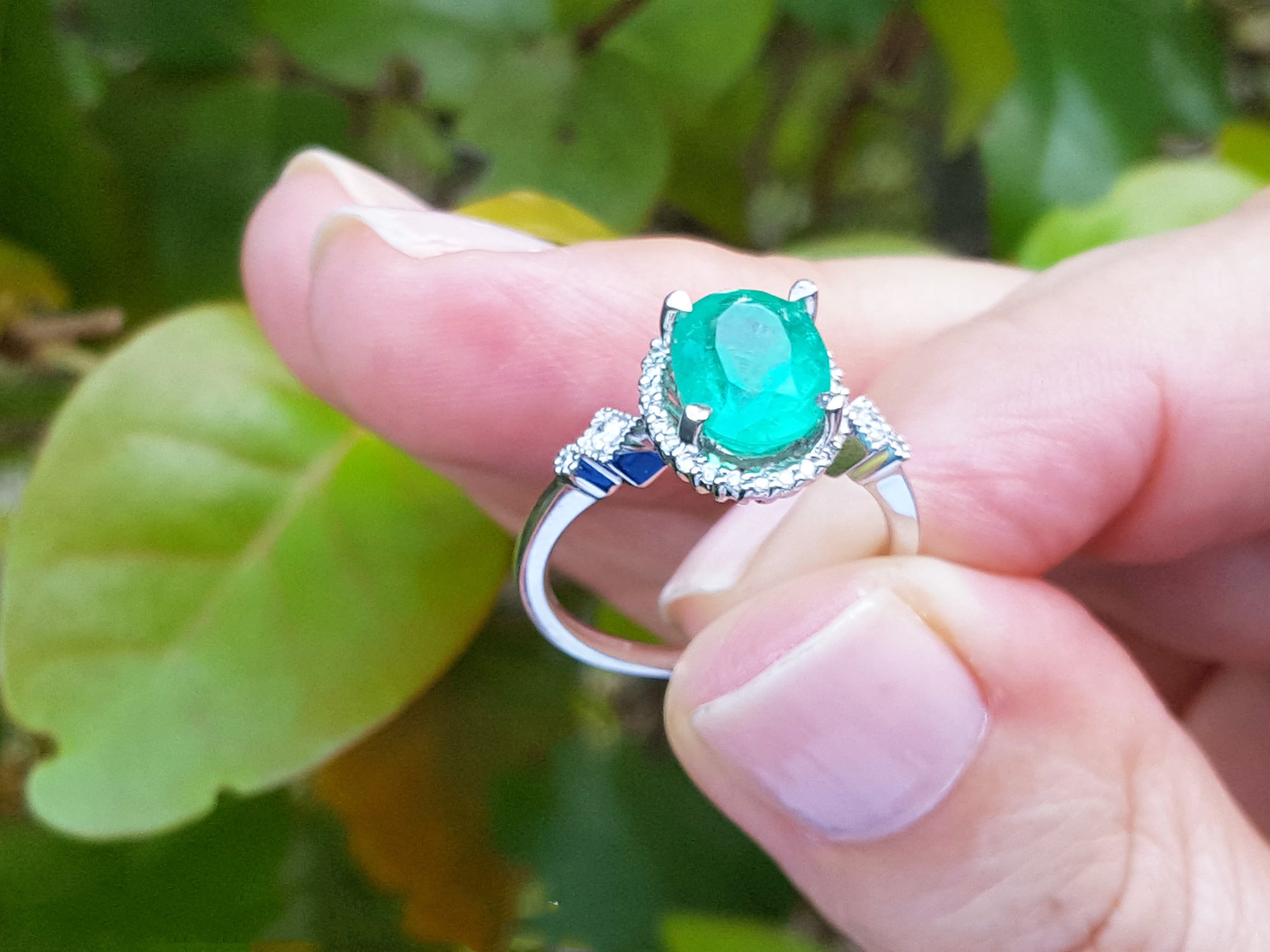 A white gold ring with n oval cut emerald held between fingers against a green leafy background.