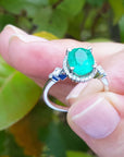 A white gold ring with n oval cut emerald held between fingers against a green leafy background.